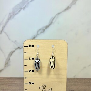 May include: A pair of dangle earrings shaped like paperclips with googly eyes, displayed on a light wood stand with a printed ruler showing a height of approximately 1.5 inches. One earring is grey and the other is a natural light wood color.