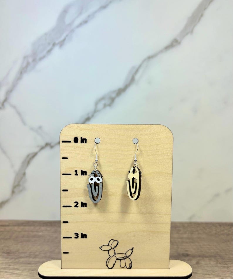 May include: A pair of dangle earrings shaped like paperclips with googly eyes, displayed on a light wood stand with a printed ruler showing a height of approximately 1.5 inches. One earring is grey and the other is a natural light wood color.