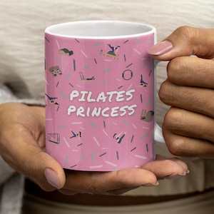 Puede incluir: Taza de cerámica blanca con fondo rosa y la frase "PILATES PRINCESS" en blanco. La taza está decorada con pequeñas ilustraciones de ejercicios de pilates y líneas rosas y azules.