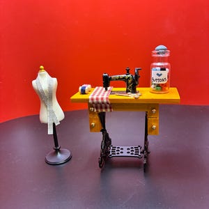 Puede incluir: Escena de costura en miniatura con una máquina de coser negra, una mesa amarilla y un tarro con la etiqueta "Buttons". También hay un maniquí con una cinta métrica y carretes de hilo. El fondo es rojo liso.
