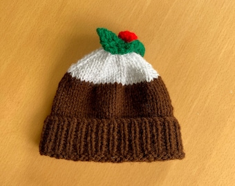 Pode incluir: Um chapéu de malha castanho com uma faixa branca e uma folha de azevinho verde com uma baga vermelha no topo. O chapéu tem a forma de um pudim de Natal.