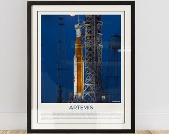 May include: Framed print featuring the Artemis rocket on its launchpad, with the NASA logo visible. The rocket is orange and white against a deep blue sky. The print is in a black frame and includes the word "ARTEMIS".