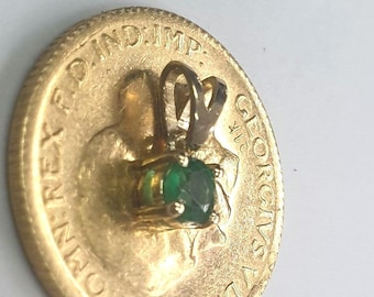 May include: A gold pendant with a vibrant green gemstone, set on a gold coin. The coin features embossed text and a profile image. The pendant's setting is silver-colored, and the gemstone is round.