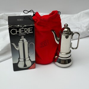May include: A silver-toned metal espresso maker with a tall cylindrical body, a curved spout, and a side handle. It is accompanied by a bright red drawstring storage bag featuring the logo 3B and a black cardboard box labelled CHÉRIE ESPRESSO.
