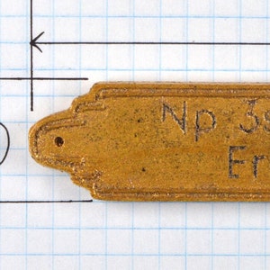 May include: A rectangular, gold-coloured plaque with decorative edges. The plaque is 148 mm long and 25 mm wide. It features the text "Np 3s font: Valentine Eric Wallis (b.1968)" engraved on its surface. Two small holes are visible.