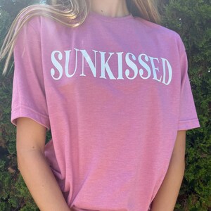 Op de afbeelding: Een roze T-shirt met het woord "SUNKISSED" in wit op de voorkant. Het shirt wordt gedragen door een persoon met lang blond haar. De persoon draagt een zwarte rok en roze en witte kralenarmbanden.