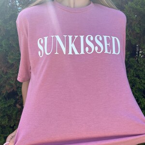 Op de afbeelding: Een mauvekleurig T-shirt met het woord "SUNKISSED" in wit op de voorkant.