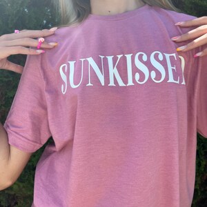Op de afbeelding: Een roze T-shirt met het woord "SUNKISSED" in wit op de voorkant.