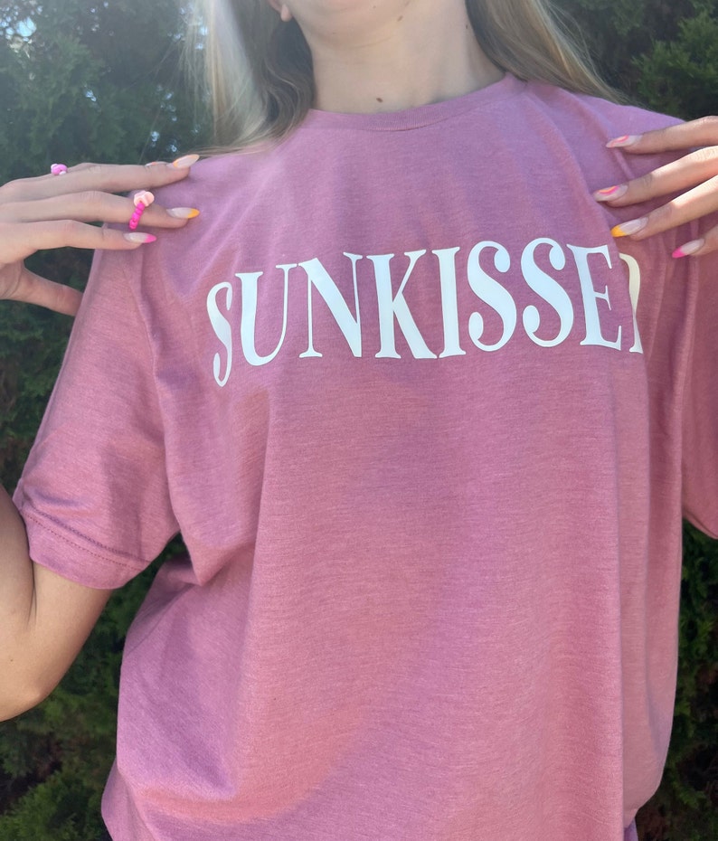 Op de afbeelding: Een roze T-shirt met het woord "SUNKISSED" in wit op de voorkant.