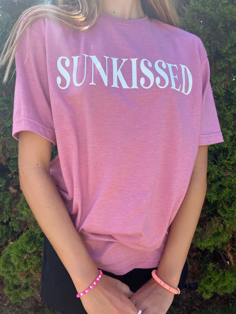 Op de afbeelding: Een roze T-shirt met het woord "SUNKISSED" in wit op de voorkant. Het shirt wordt gedragen door een persoon met lang blond haar. De persoon draagt een zwarte rok en roze en witte kralenarmbanden.