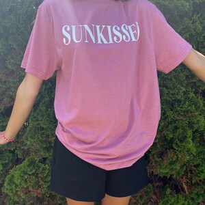 Op de afbeelding: Een roze T-shirt met het woord "SUNKISSED" in wit op de voorkant. Het shirt wordt gedragen door een persoon en wordt gecombineerd met een zwarte short.