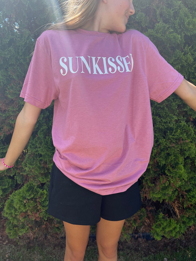 Op de afbeelding: Een roze T-shirt met het woord "SUNKISSED" in wit op de voorkant. Het shirt wordt gedragen door een persoon en wordt gecombineerd met een zwarte short.