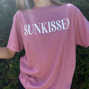 Op de afbeelding: Een roze T-shirt met het woord "SUNKISSED" in wit op de voorkant.