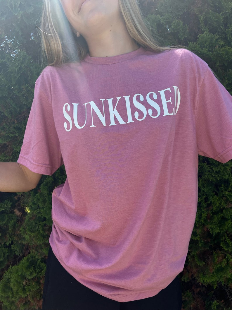 Op de afbeelding: Een roze T-shirt met het woord "SUNKISSED" in wit op de voorkant.