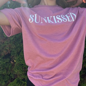 Op de afbeelding: Een roze T-shirt met het woord "SUNKISSED" in wit op de voorkant.