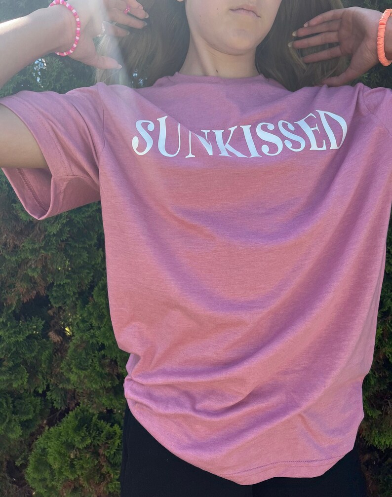 Op de afbeelding: Een roze T-shirt met het woord "SUNKISSED" in wit op de voorkant.