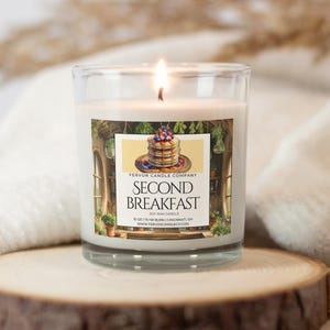 May include: A lit soy wax candle in a clear glass jar. The label features an illustration of pancakes with berries and the text "SECOND BREAKFAST". The candle is on a wooden surface.