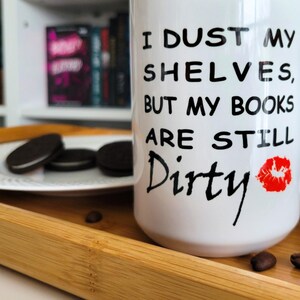 White and black coffee mug with a playful book lover's quote: "I dust my shelves, but my books are still dirty," featuring a red lipstick kiss mark. The mug is placed on a wooden tray with cookies and coffee beans, and a bookshelf filled with romantasy and dark romance books in the background. A perfect gift for bookworms, readers, and bibliophiles with a sense of humor. Ideal for cozy reading sessions and literary-themed gifts.