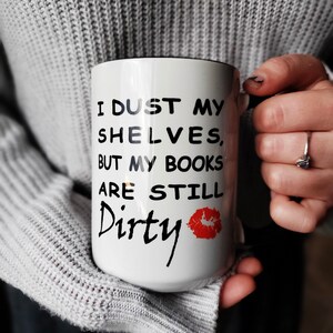 White coffee mug with black text that reads 'I dust my shelves, but my books are still dirty' with a red lipstick kiss mark.