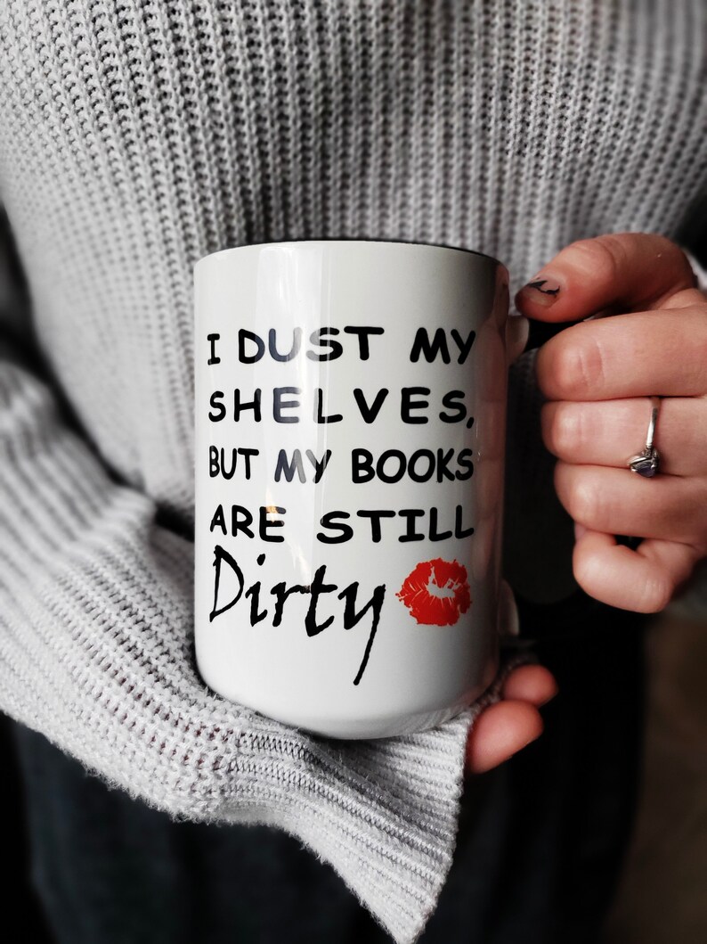 White coffee mug with black text that reads 'I dust my shelves, but my books are still dirty' with a red lipstick kiss mark.