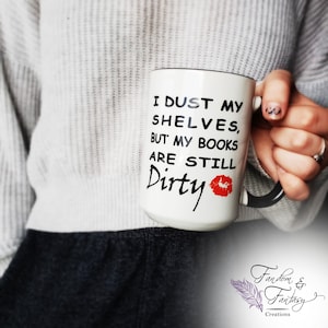 White ceramic coffee mug with black text that reads 'I dust my shelves, but my books are still Dirty' with a red lipstick kiss mark. The mug has a black handle.