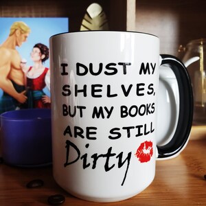 White coffee mug with a black handle and rim. The mug has black text that reads 'I dust my shelves, but my books are still dirty' with a red lipstick kiss mark next to the word dirty. It sits on a wooden tray with bookish fan art in the background of April and Mitch from Jen DeLuca's book, Well Matched.