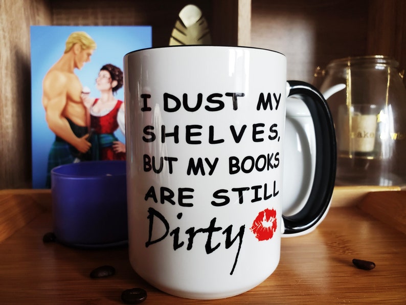 White coffee mug with a black handle and rim. The mug has black text that reads 'I dust my shelves, but my books are still dirty' with a red lipstick kiss mark next to the word dirty. It sits on a wooden tray with bookish fan art in the background of April and Mitch from Jen DeLuca's book, Well Matched.