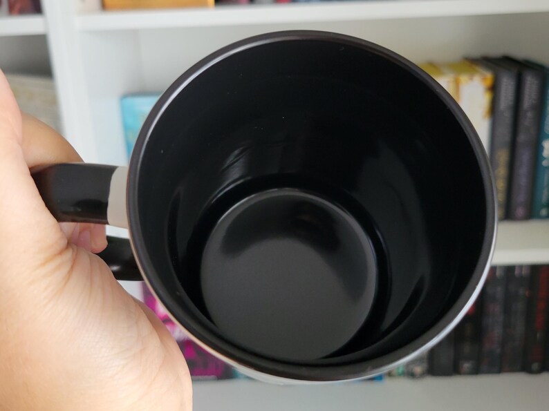 **ALT Text for Etsy Listing Image:**  
Close-up of a black-handled coffee mug with a glossy black interior, held by a hand against a bookshelf filled with romantasy and dark romance books. The smooth ceramic finish highlights the high-quality craftsmanship. Perfect for book lovers, fantasy readers, and coffee enthusiasts looking for a stylish and unique mug. Ideal for gifting or personal use.