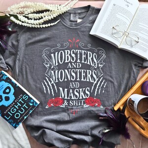 May include: Grey t-shirt with the text "Mobsters and Monsters and Masks & Shit" in white, red, and black. The shirt features roses, guns, and claw marks. A book and open book are also in the image.