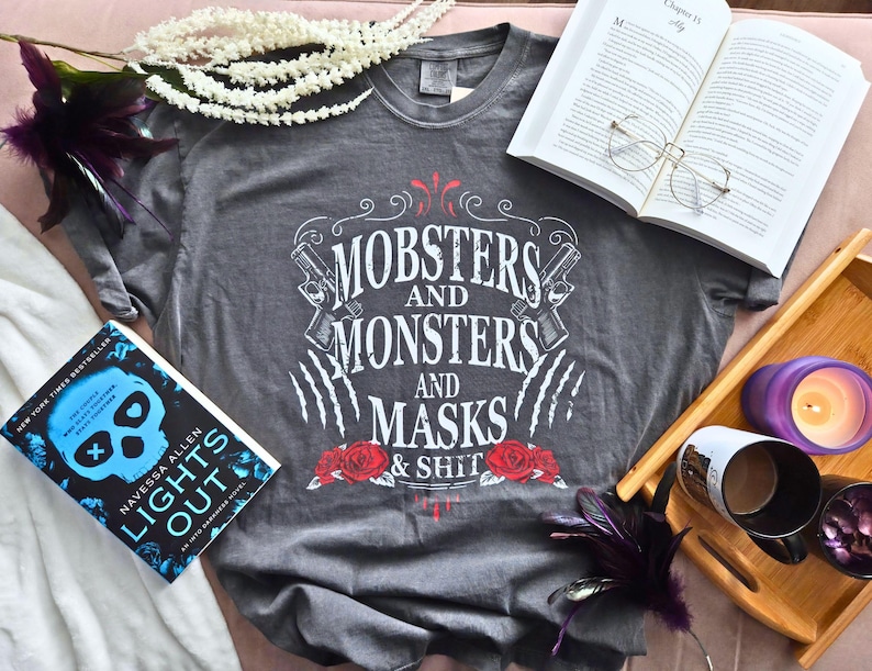 May include: Grey t-shirt with the text "Mobsters and Monsters and Masks & Shit" in white, red, and black. The shirt features roses, guns, and claw marks. A book and open book are also in the image.