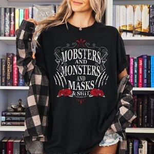 May include: Black t-shirt featuring the text "MOBSTERS AND MONSTERS AND MASKS & SHIT" in white, with a design of guns, claws, and roses. The person is wearing a black and white checked shirt over the t-shirt.