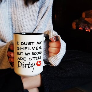 White coffee mug with a black handle and rim and black text that reads 'I dust my shelves, but my books are still dirty' with a red lipstick kiss mark.