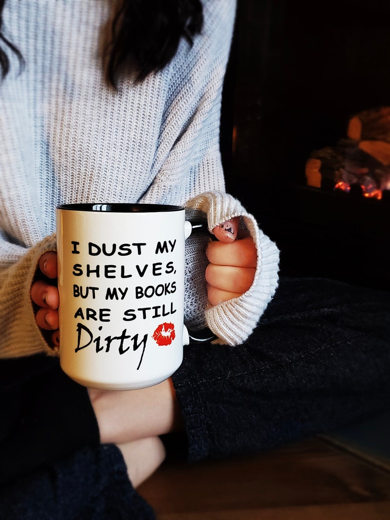 White coffee mug with a black handle and rim and black text that reads 'I dust my shelves, but my books are still dirty' with a red lipstick kiss mark.
