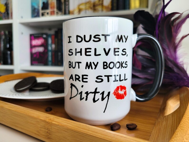 White and black coffee mug with a playful book lover's quote: "I dust my shelves, but my books are still dirty," featuring a red lipstick kiss mark. The mug is placed on a wooden tray with cookies and coffee beans, and a bookshelf filled with romantasy and dark romance books in the background. A perfect gift for bookworms, readers, and bibliophiles with a sense of humor. Ideal for cozy reading sessions and literary-themed gifts.