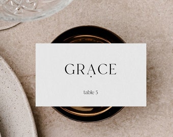 May include: A white place card with the name "GRACE" and the text "table 5" printed in black. The place card is sitting on a brown coaster.