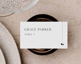 May include: A white place card with black text that reads "GRACE PARKER TABLE 5" and a small leaf design in the bottom right corner. The place card is sitting on a brown surface.