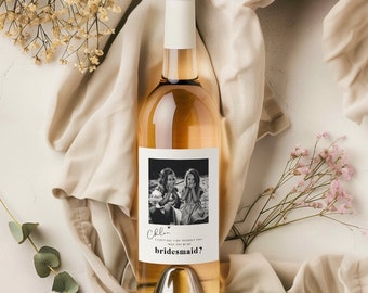 May include: A bottle of white wine with a black and white photo of two girls on the label. The label says "Cheers... I can't say I'd  without you! Will you be my bridesmaid?"