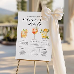 May include: A white wedding sign on a gold easel, featuring the text "SIGNATURE drinks." The sign displays illustrations of three cocktails with their ingredients listed below. A cream-colored bow and fabric drape the side.