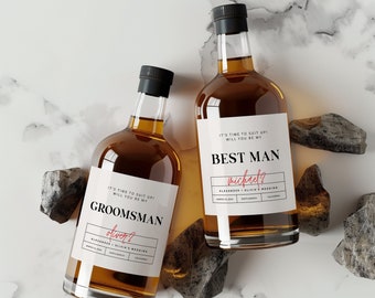 May include: Two glass bottles of whiskey with labels that say "It's time to suit up! Will you be my Best Man?" and "It's time to suit up! Will you be my Groomman?" The labels are white with black text and red accents. The bottles are sitting on a white marble surface with gray rocks.