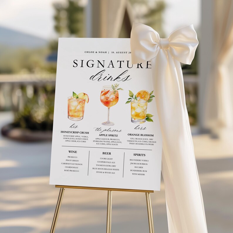 May include: A white wedding sign on a gold easel, featuring the text "SIGNATURE drinks." The sign displays illustrations of three cocktails with their ingredients listed below. A cream-colored bow and fabric drape the side.