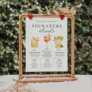 May include: A copper-colored metal frame holds a white sign with the words "SIGNATURE drinks." The sign features illustrations of three cocktails and lists ingredients. Dried flowers and red velvet accents adorn the top of the frame. The sign is for a wedding.