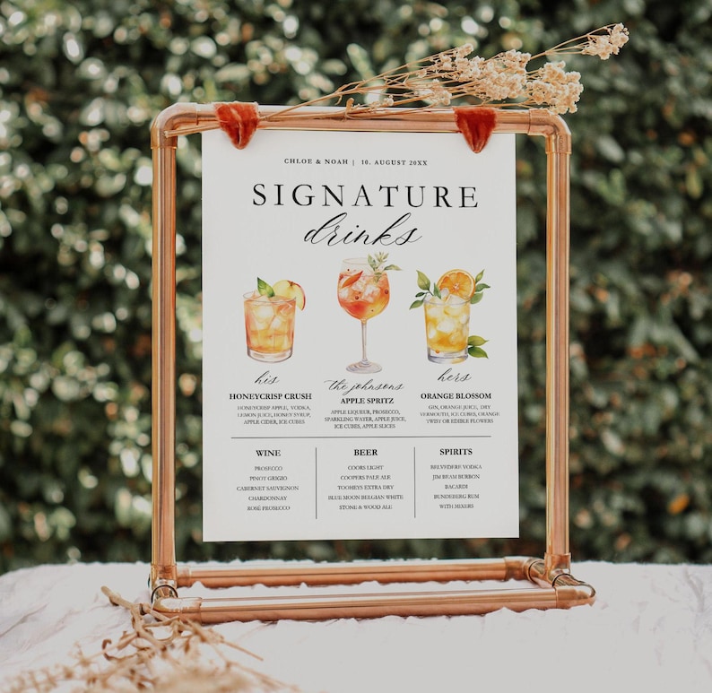 May include: A copper-colored metal frame holds a white sign with the words "SIGNATURE drinks." The sign features illustrations of three cocktails and lists ingredients. Dried flowers and red velvet accents adorn the top of the frame. The sign is for a wedding.