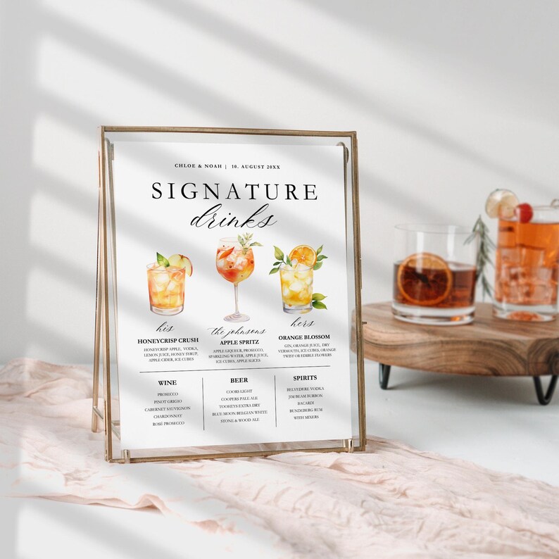 May include: A framed wedding sign with a gold metal frame displays the words "Signature Drinks." The sign features illustrations of three cocktails: Honeycrisp Crush, Apple Spritz, and Orange Blossom. The sign also lists wine, beer, and spirits.