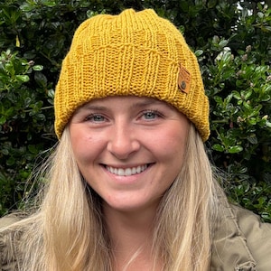 May include: A mustard yellow knit beanie featuring a vertical ribbed texture and a folded brim. A small, rectangular brown leather tag is attached to the side of the beanie, secured with a single metal rivet, adding a subtle detail to the design.