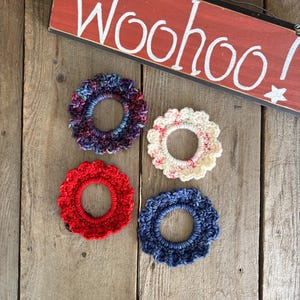 May include: Four circular crochet hair scrunchies in variegated purple, solid red, cream with pink speckles, and solid navy blue. Above them is a rectangular red wooden sign with the white painted text "Woohoo!" and a small white star.