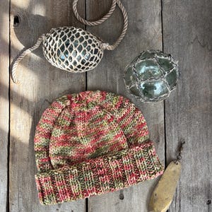 May include: A knitted hat with a red, green, and brown color pattern. The hat is sitting on a wooden surface with a fishing lure, a glass buoy, and a rope.