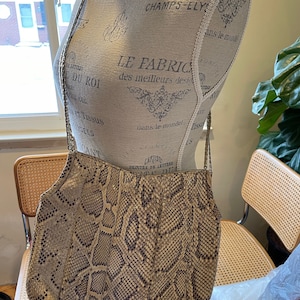 May include: A beige snakeskin shoulder bag with a brown strap. The bag is on a mannequin with a printed fabric covering.