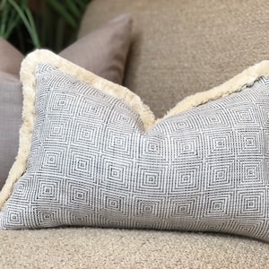 May include: A grey and white patterned cushion with a cream-coloured fringe border. The cushion is sitting on a beige sofa.