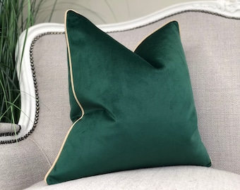 May include: A dark green velvet pillow with a cream colored border. The pillow is sitting on a light gray couch.