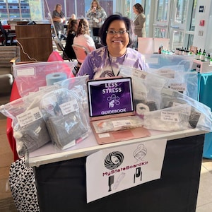 May include: A pink laptop with a black screen displaying the text "LESS STRESS GUIDEBOOK". The laptop is on a table with various items in clear plastic bags, including a black and gray pouch, a white towel, and a black and white pouch. The table has a black tablecloth with a white and black geometric pattern. A sign with the text "MyStateBoardKit" is on the table. The sign features a black and white illustration of a person with a towel wrapped around their head.
